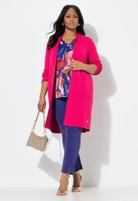 Long cardigan rose vif sur un haut à motifs floraux bleu, orange et rose. Associé à un pantalon marine et des sandales dorées, tenant un sac à main pailleté.