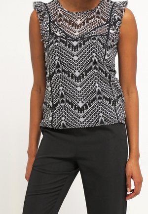 Hihaton pusero, jossa mustavalkoinen geometrinen kuvio ja röyhelöidyt olkapäät, yhdistetty mustiin istuviin housuihin seisovalla henkilöllä.