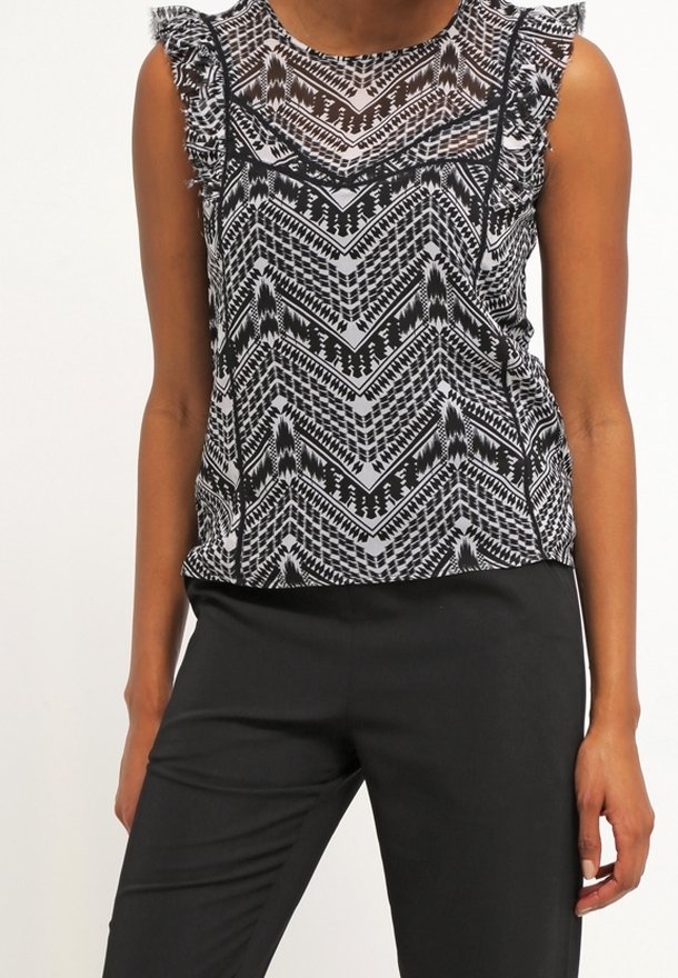Blouse sans manches à motif géométrique noir et blanc avec épaules à volants, portée avec un pantalon noir ajusté sur une personne debout.