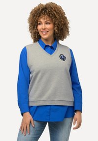Grå strikket vest med V-halsdesign, der har en blå skjorte under. Vesten inkluderer et broderet logo på brystet.