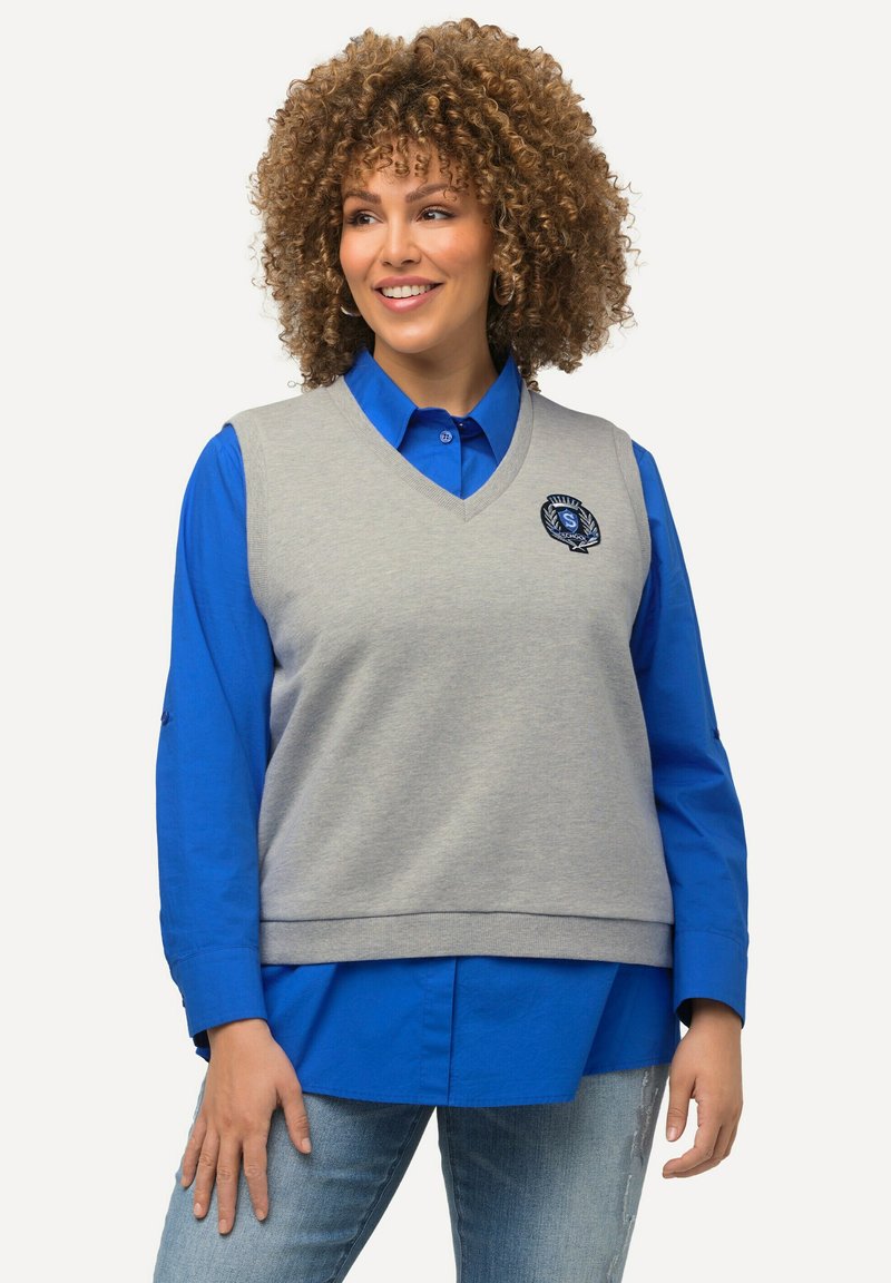 Grå strikket vest med V-halsdesign, der har en blå skjorte under. Vesten inkluderer et broderet logo på brystet.