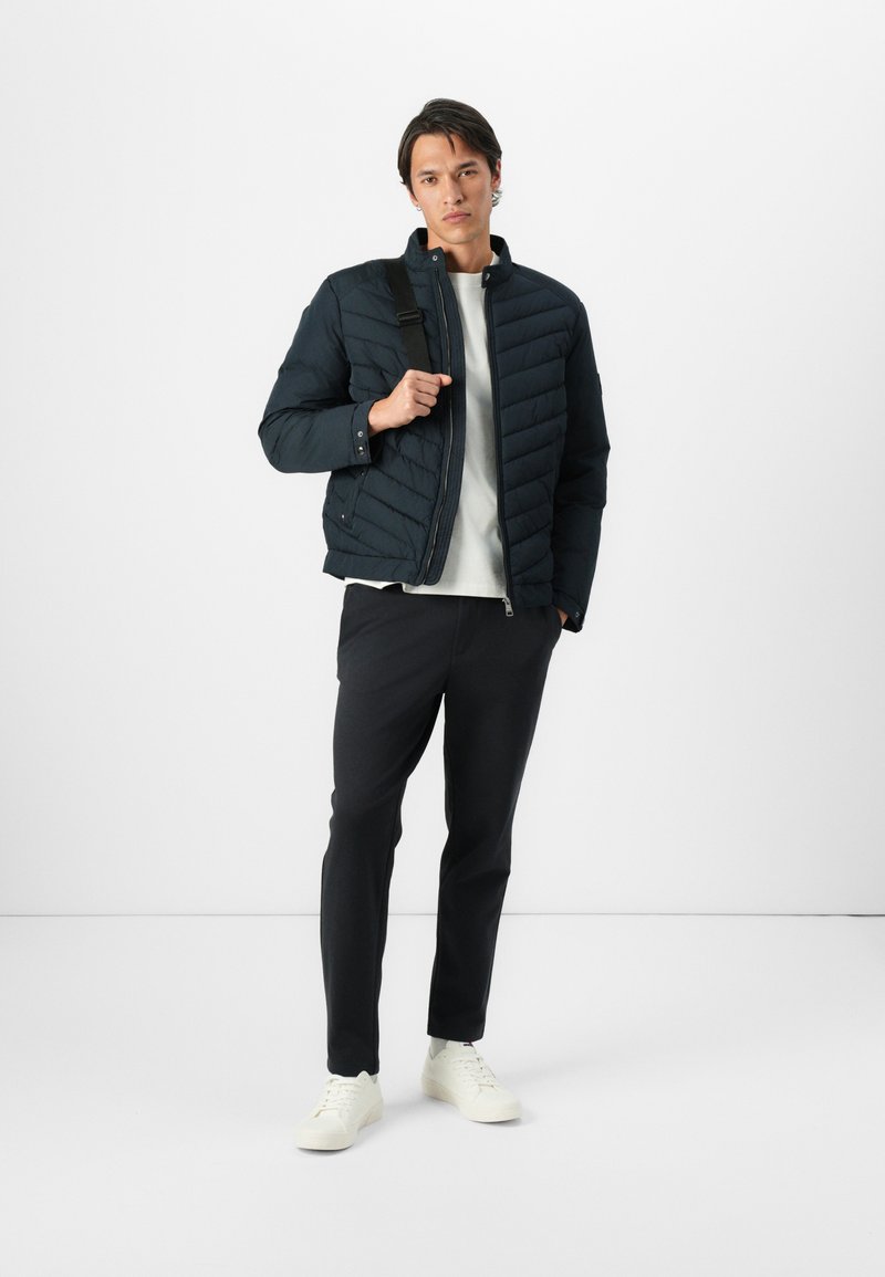 Mężczyzna w czarnej pikowanej kurtce, białej koszuli, czarnych spodniach, białych adidaskach i noszący czarną torbę na ramię na białym tle.