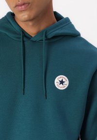 Tyrkise fleece hettegenser med snor, med en rund hvit lapp med rød og blå tekst "CONVERSE ALL STAR" og et stjernedesign.