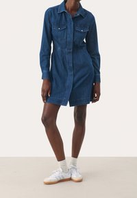 Denim-mekko, jossa on kapea vyötärö, pitkät hihat, rintataskut ja nappikiinnitys. Tumansinisä väri ja klassinen kaulus.