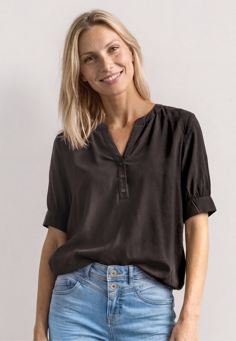 Femme souriante aux cheveux blonds portant un chemisier noir boutonné et un jean bleu clair sur un fond uni.