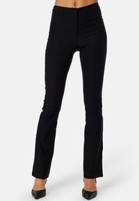 Zwarte slim-fit broek met een gladde textuur, hoge taille en subtiele details, gecombineerd met puntige zwarte hakken.
