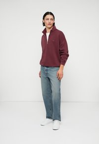 Un jeune homme portant un sweat à col zippé bordeaux, un jean bleu clair et des baskets blanches, se tenant sur un fond blanc uni.