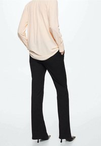 Langärmlige Bluse in Hellpeach mit lockerer Passform, kombiniert mit eng anliegenden schwarzen Hosen und spitzen schwarzen Absätzen, von hinten gesehen.