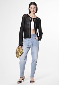 Schwarze Netzstrickjacke mit weißen Akzenten, kombiniert mit hochgeschnittenen, hellblauen Jeans und schwarzen Sandaletten mit offenen Zehen, während sie eine goldene, texturierte Clutch hält.
