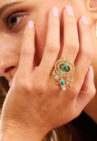 Gold ring featuring a large green gemstone, surrounded by smaller colored gems. Intricate texture and design with dangling accents.