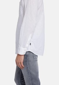 Chemise en lin blanche à manches longues avec des poignets à boutons, une poche poitrine unique et une texture subtile, associée à un jean gris ajusté.