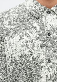 Juoda ir balta tekstūruota marškinėlių palaidinė su apykakle ir juodais mygtukais, visame paviršiuje subtilus abstraktus raštas.