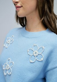 Suéter azul claro con cuello redondo, que presenta tres diseños de flores blancas bordadas adornadas con acentos de perlas. Textura suave.