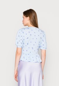 Top peplum azul claro com mangas bufantes, apresentando um padrão floral. O tecido tem uma textura suave, complementado por uma saia lilás lisa.
