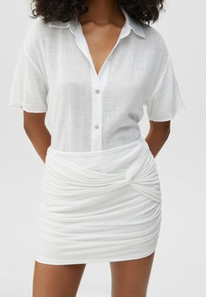 Femme portant une chemise blanche à manches courtes boutonnée, rentrée dans une mini-jupe blanche froncée avec un détail de nœud torsadé à la taille.