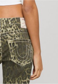 Zielone dżinsy w lamparcim wzorze z białym przeszyciem, z emblematem na tylnej kieszeni i metką marki na pasku. 