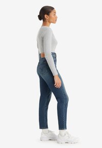 Jeans de ganga de cintura alta em azul médio com perna ajustada. Combinados com uma blusa de manga longa canelada cinza clara e ténis brancos.