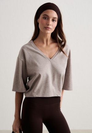 Donna che indossa una maglietta beige a maniche corte con scollo a V, leggings marrone scuro e una fascia larga coordinata, in piedi davanti a uno sfondo semplice.