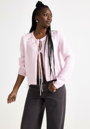 Ljusrosa stickad kofta med knytband fram, dekorerad med pärldetaljer på axlarna och ribbade muddar, kombinerad med mörka jeans.