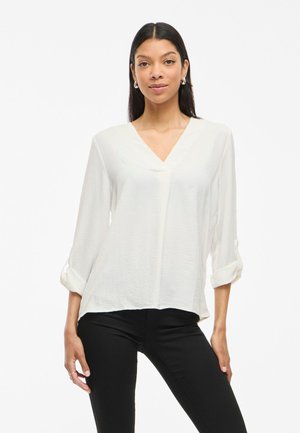 Weißes V-Ausschnitt-Bluse mit langen Ärmeln, die an den Manschetten hochgerollt sind, aus leichtem Stoff gefertigt, mit entspannter Passform und subtiler Textur. Kombiniert mit schwarzen Hosen.