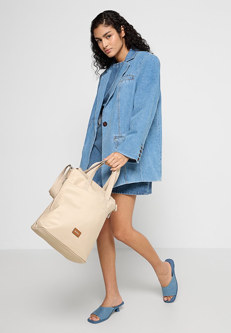 Frau trägt einen Jeansblazer und einen Rock mit passenden blauen Schuhen und hält eine große beige Tote-Tasche, während sie vor einem schlichten weißen Hintergrund geht.