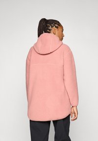 Eine Person mit geflochtenem Haar blickt abgewandt und trägt einen weichen, rosa Kapuzenpullover. Der Hintergrund ist schlicht weiß, was die Textur des Kleidungsstücks hervorhebt.