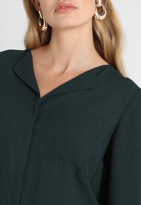 Blusa verde scuro con colletto classico, tasca frontale e texture liscia, abbinata a grandi orecchini a cerchio dorati.
