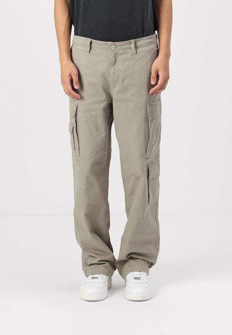 Beige cargo bukser med en afslappet pasform, der har to sidelommer, en knaplukning og kontrastsyning. Paret med hvide sneakers.