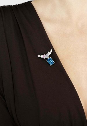 Silver brooch with blue gemstone, pearl, and pink accents, featuring a wing design. Set against a dark fabric background.