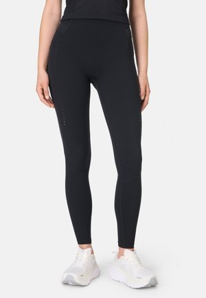 Vrouw draagt zwarte strakke leggings en witte sportschoenen, staand tegen een effen lichte achtergrond.