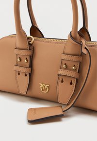 Tan leather handbag with gold hardware, dual top handles, accentuated stitching, and a logo detail. Includes a decorative tag attached by a strap.