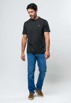 Vīrietis iet, valkājot melnu īsroku T-kreklu, zilas džinsa bikses un brūnas sporta kurpes, vienkāršas gaiši pelēkas fona priekšā.