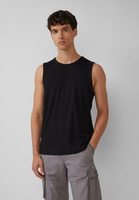 Schwarzes ärmelloses Tanktop aus glattem Stoff, mit einem runden Ausschnitt und einer figurbetonten Silhouette. Kombiniert mit grauen Cargo-Shorts.