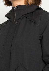 Černá bunda s vysokým límcem, zipovým zapínáním a patentkami. Obsahuje strukturovanou látku a jemné akcenty na ramenou.