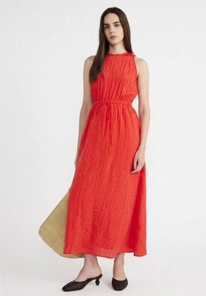 Femme en robe longue rouge sans manches cintrée à la taille, tenant un grand sac fourre-tout tressé et portant des chaussures tressées noires à talons plats.