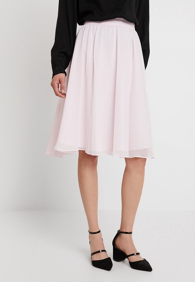 Falda corta de color rosa claro, hasta la rodilla, hecha de un tejido transparente con una textura suave, que presenta una cinturilla elástica y un diseño de capas. Un par de zapatos de tacón negros.