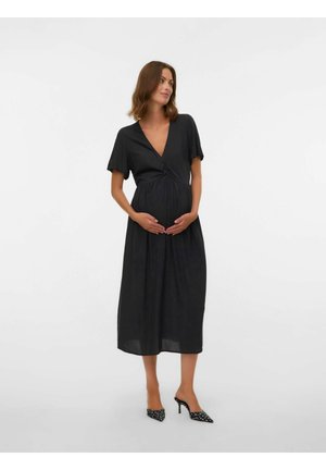 Femme enceinte debout dans une robe midi noire à manches courtes et col en V profond, portant des escarpins noirs à pois, les mains sur le ventre.
