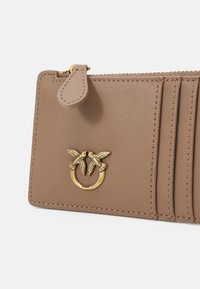 Carteira de couro castanho com emblema de pássaro em tom dourado, fecho de zip, múltiplos compartimentos para cartões e uma superfície texturizada.