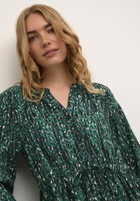 Bluză verde cu model, cu accente negre și crem, având guler, nasturi și o croială lejeră cu un șnur la talie.