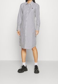 Donna che indossa un abito bianco a maniche lunghe e lunghezza ginocchio, con righe verticali nere, scarpe nere chunky e calzini bianchi su uno sfondo semplice.