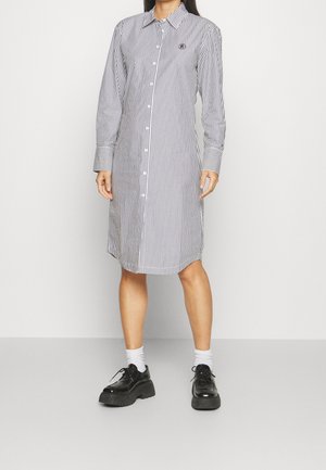 Frau in einem knielangen, langärmligen, weißen Kleid mit schwarzen vertikalen Streifen, schwarzen klobigen Schuhen und weißen Socken vor einem schlichten Hintergrund.