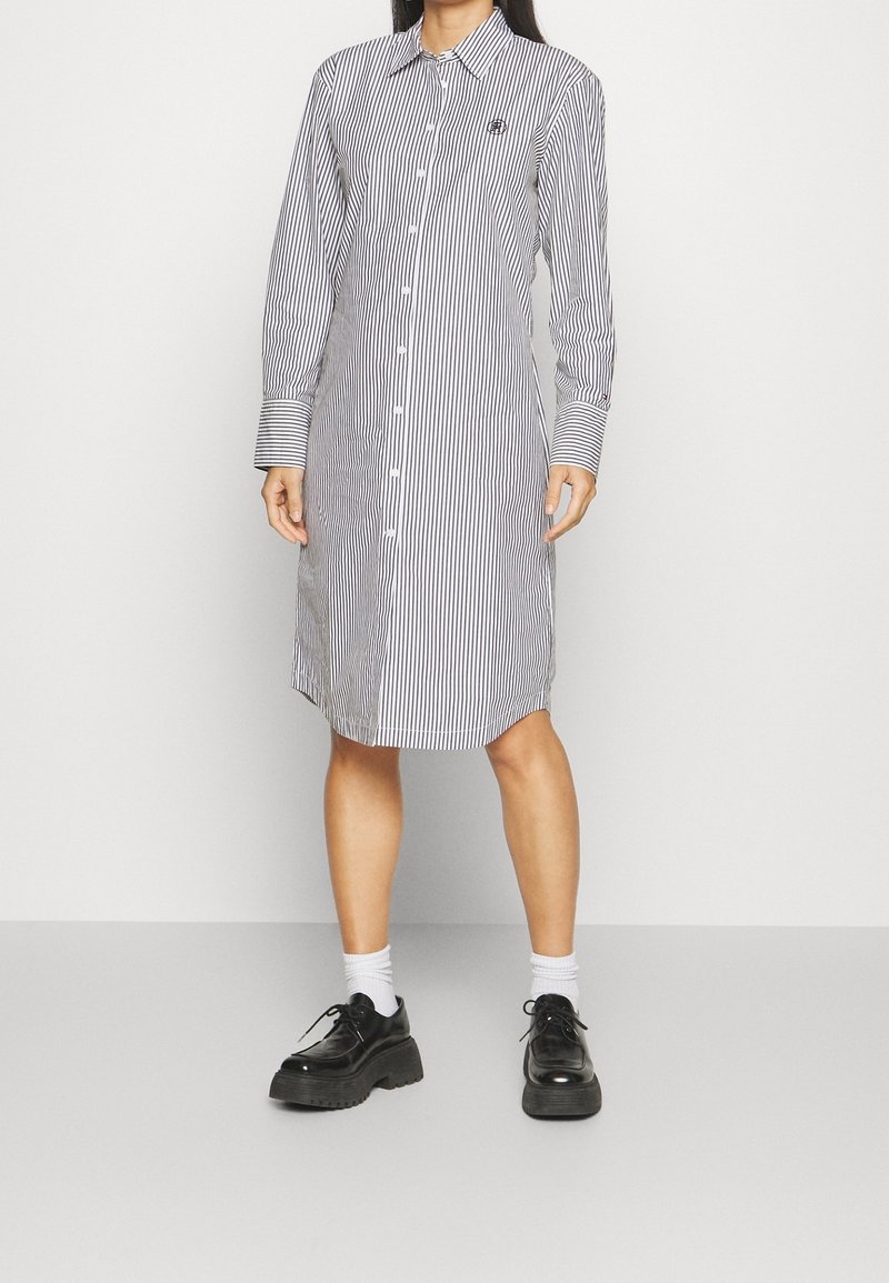 Donna che indossa un abito bianco a maniche lunghe e lunghezza ginocchio, con righe verticali nere, scarpe nere chunky e calzini bianchi su uno sfondo semplice.