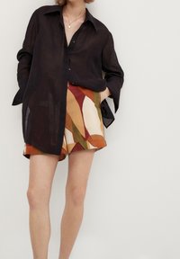 Osoba w przejrzystej, czarnej koszuli z długim rękawem oraz szortach w abstrakcyjne wzory w ziemistych odcieniach, stoi z rękami w kieszeniach na jednolitym tle.