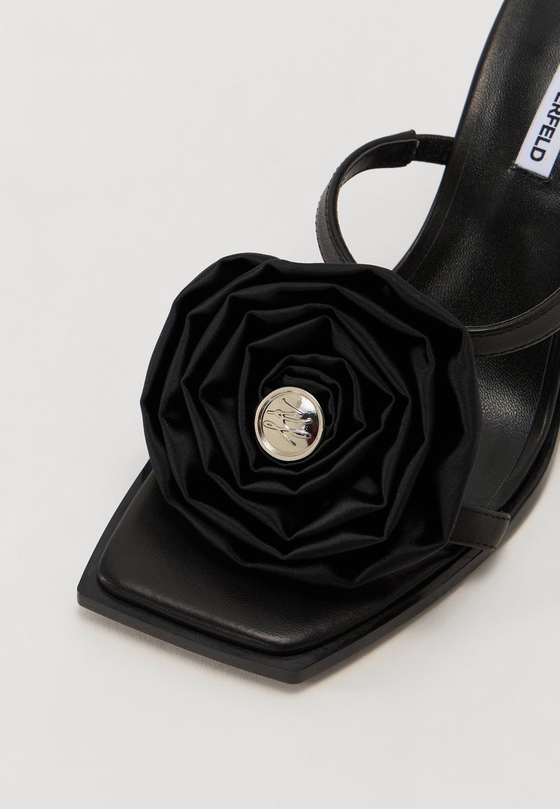 Schwarze High-Heel-Sandalette mit quadratischer Zehenpartie, versehen mit einem strukturierten Satinblütenakzent und einer silbernen Logo-Verzierung in der Mitte.