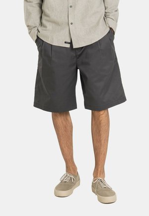 Man wearing beige button-up shirt, dark gray pleated shorts, and light beige lace-up sneakers, standing with hands in pockets.