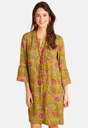 Frau lächelt, trägt ein grünes Blumenkleid mit 3/4-Ärmeln, kombiniert mit einer langen roten Perlenkette und passenden roten Ohrringen.