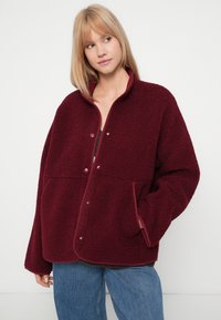 Blond naine, kes kannab suure suurusega burgundia värvi fliisjakki, millel on nööbid ja siniseid teksaseid, seismas halli valge tausta ees.