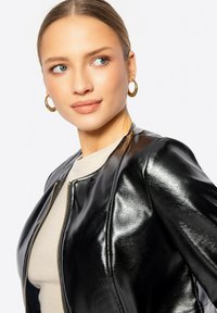 Giubbotto bomber in pelle nera con una texture liscia, colletto rotondo e dettagli in zip dorata, abbinato a un top beige sottostante.