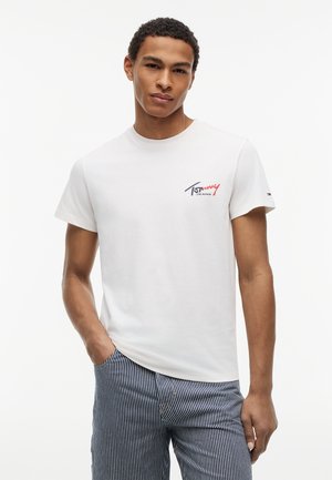 Jonge man in een wit Tommy Jeans T-shirt en blauw-witte gestreepte broek, staand met één hand in de zak tegen een effen achtergrond.