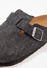Tumma harmaa huopainen liukupohjainen kenkä, jossa on teksturoitu päällinen, leveä remmi ja metallinen solki. Pehmeä pohjallinen ja kumipohja.